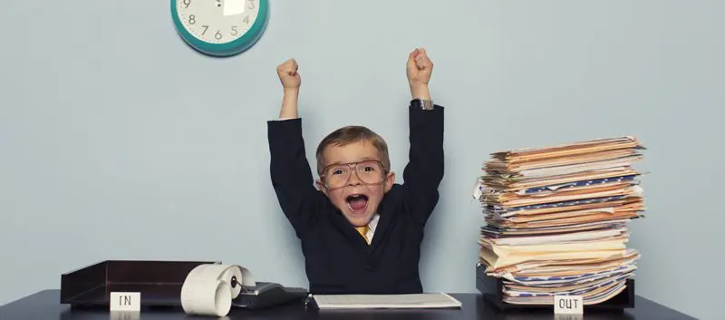 Small boy wearing a suit, sitting at a desk, throwing his arms up in the air, with an empty 'In' tray, and a full 'Out' tray.