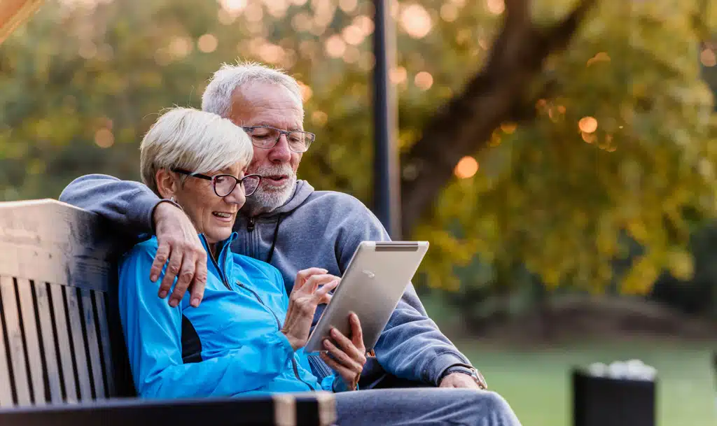 Elderly man and woman sat on a bench looking at an electronic tablet.