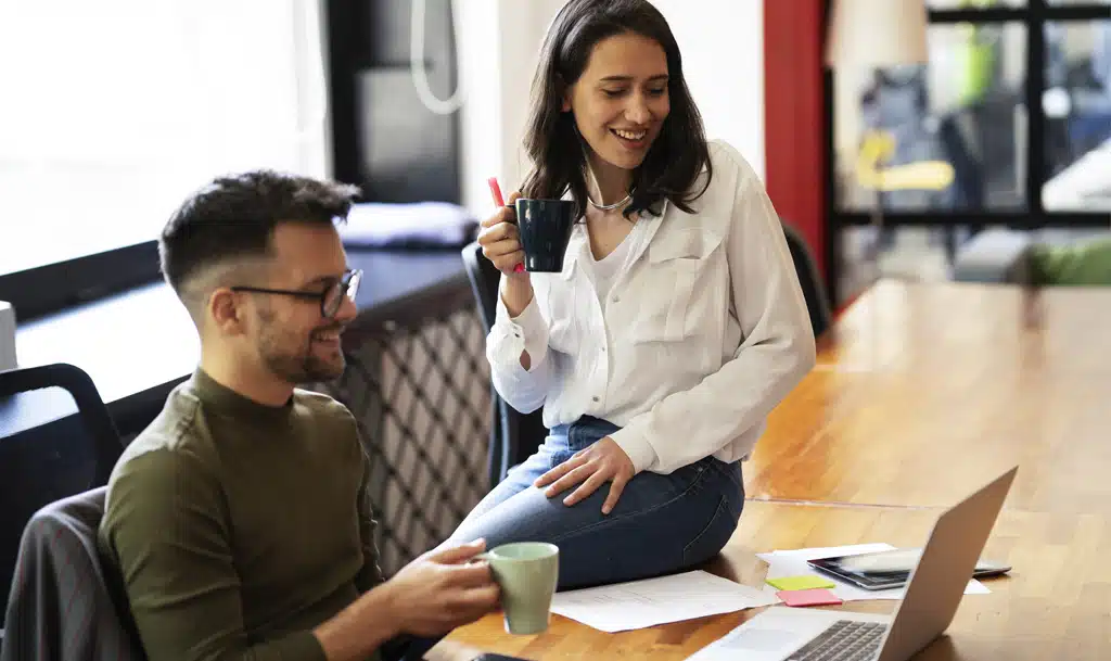 A male and female colleague sitting at a desk, drinking coffee, talking about data.