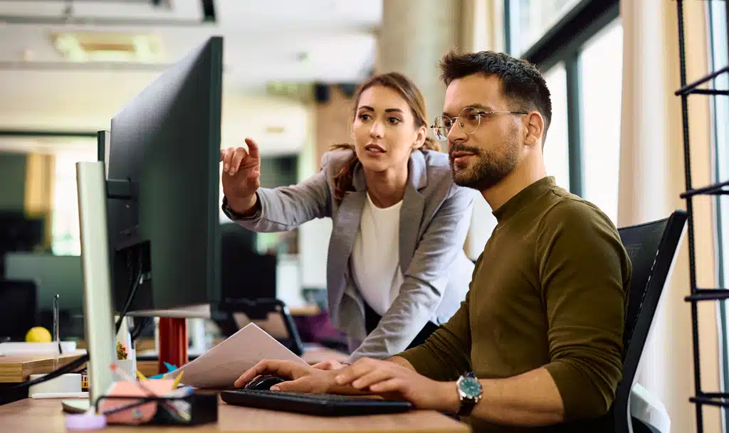 Young man sitting at a desk looking at a monitor while a young woman standing next to him points at something on the screen.