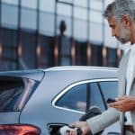 A grey-haired man in a light grey suit is plugging in the charger to his electric vehicle while holding his phone.