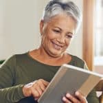 An older women is sat on a sofa looking a tablet with a smile on her face.